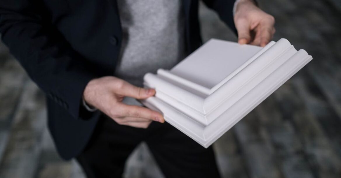 Person holding a white decorative cornice or molding sample, wearing a dark blazer and gray sweater.