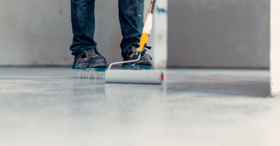 Close-up of a worker wearing spiked shoes using a paint roller to apply a coating to a smooth concrete floor during renovation