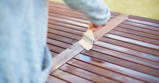 Person applying varnish or wood stain to a wooden surface using a paintbrush.