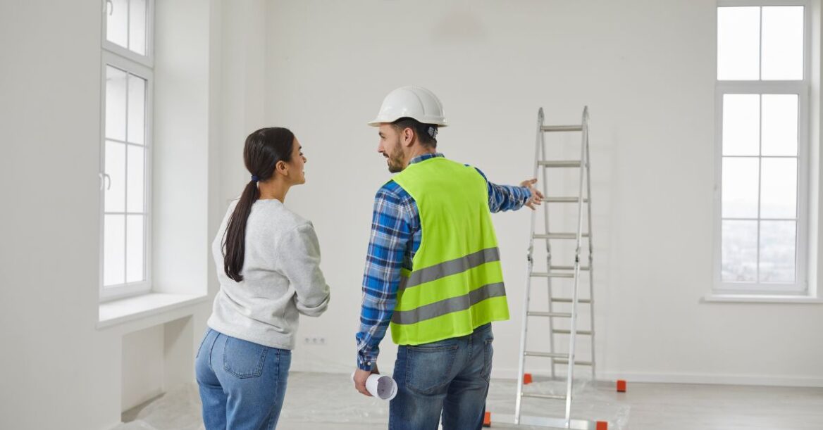 A contractor in a safety vest and hard hat pointing while speaking with a homeowner in an empty room prepared for renovation.