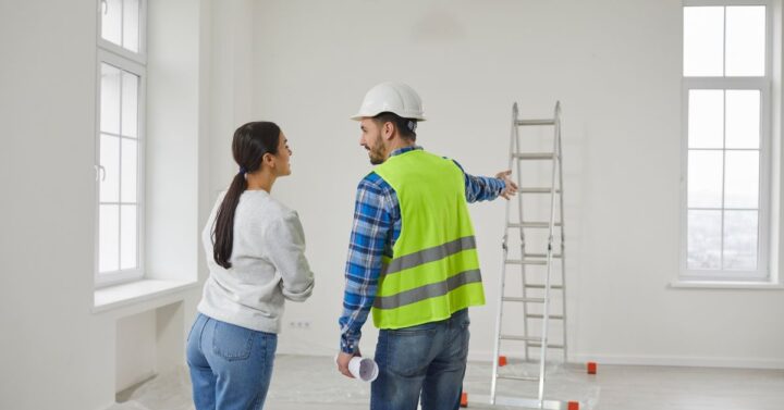 A contractor in a safety vest and hard hat pointing while speaking with a homeowner in an empty room prepared for renovation.