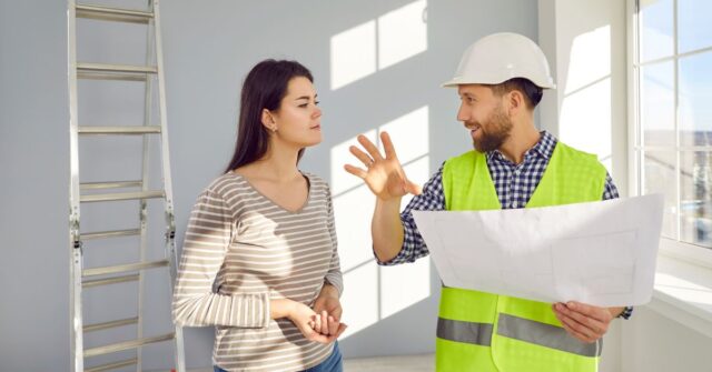 A construction worker wearing a hard hat and safety vest explains renovation plans on a blueprint to a woman inside a bright room, with a ladder and window in the background.