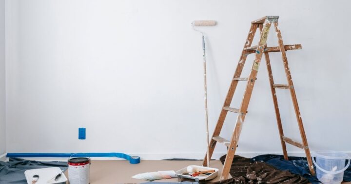 A room in the middle of a DIY painting project with a wooden ladder, paint roller, open paint can, and various painting supplies set against a white wall.