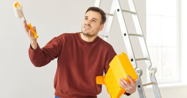 A man holding a paint roller and tray looks frustrated while painting a wall, with a ladder behind him in a bright room.