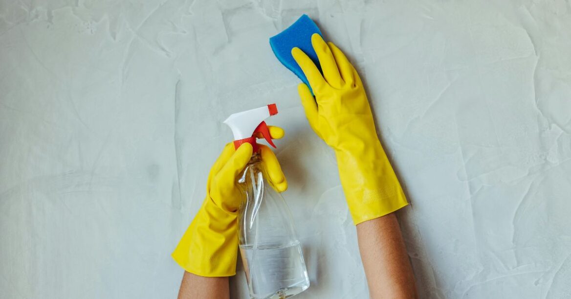 Person wearing yellow rubber gloves cleaning a wall with a spray bottle and a blue sponge.