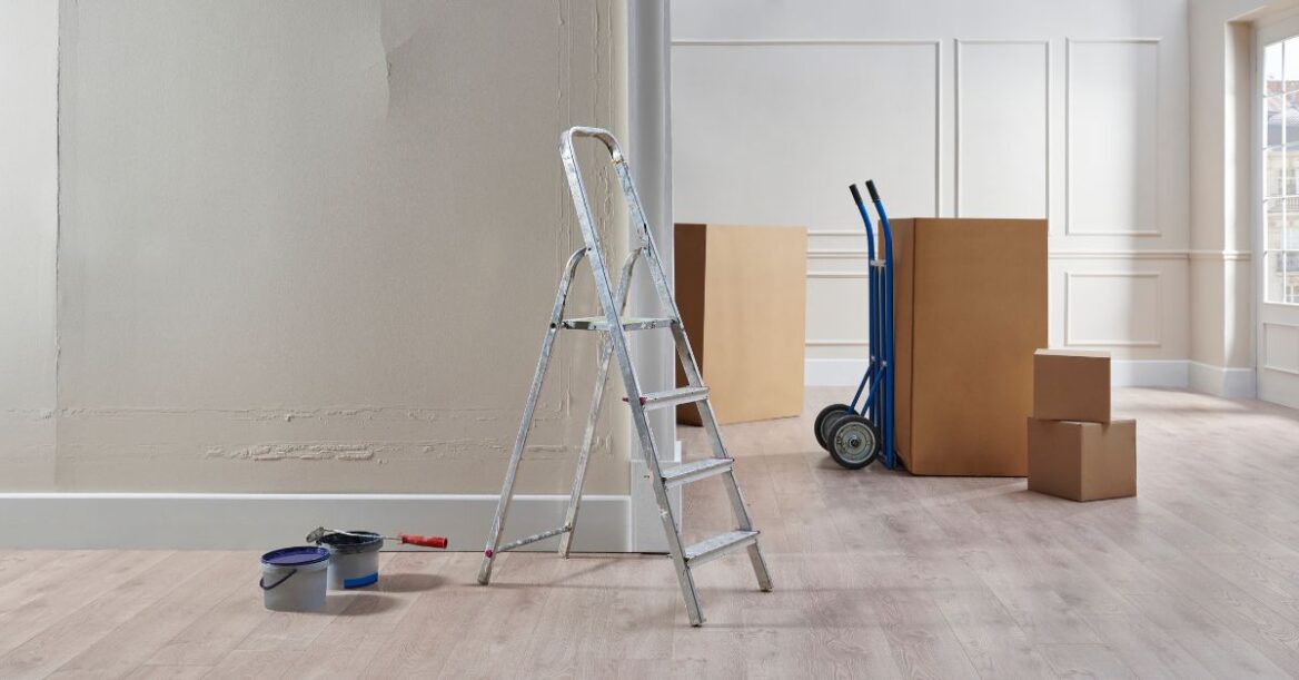 Ladder, paint supplies, moving boxes, and hand truck inside a room under renovation.