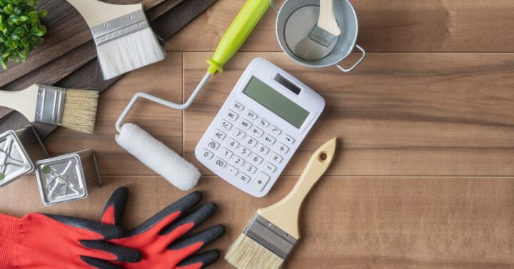 Overhead view of painting supplies on a wooden surface, including paintbrushes, a roller, gloves, paint cans, a small bucket, and a calculator, suggesting planning or estimating costs for a home painting project.