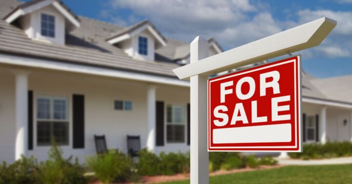 A red “For Sale” sign in front of a suburban home with a porch and lawn under a blue sky.