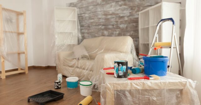 A living room with furniture covered in plastic, paint cans, rollers, brushes, and a ladder set up for a home painting project.