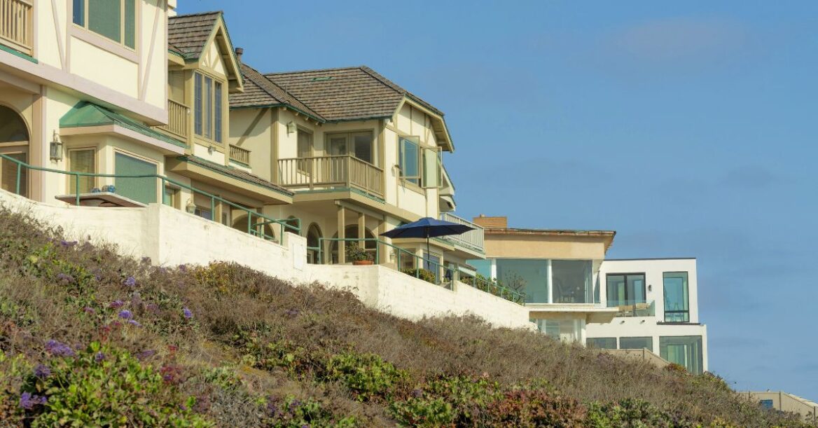 Luxury beachfront homes on a landscaped hillside overlooking a rocky shoreline beneath a clear sky.