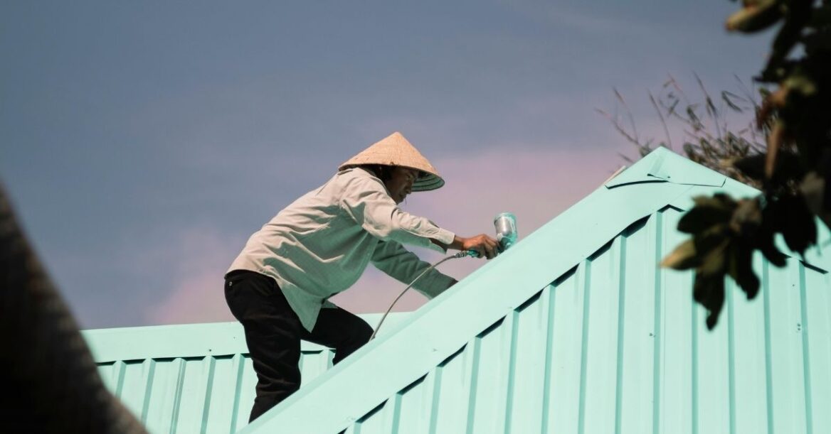 Worker in conical hat spray painting a metal roof light blue — professional roof painting and maintenance under clear sky.