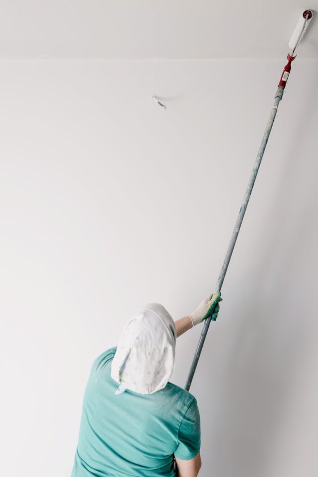 Person wearing a headscarf and gloves painting a ceiling with a roller attached to a long pole.