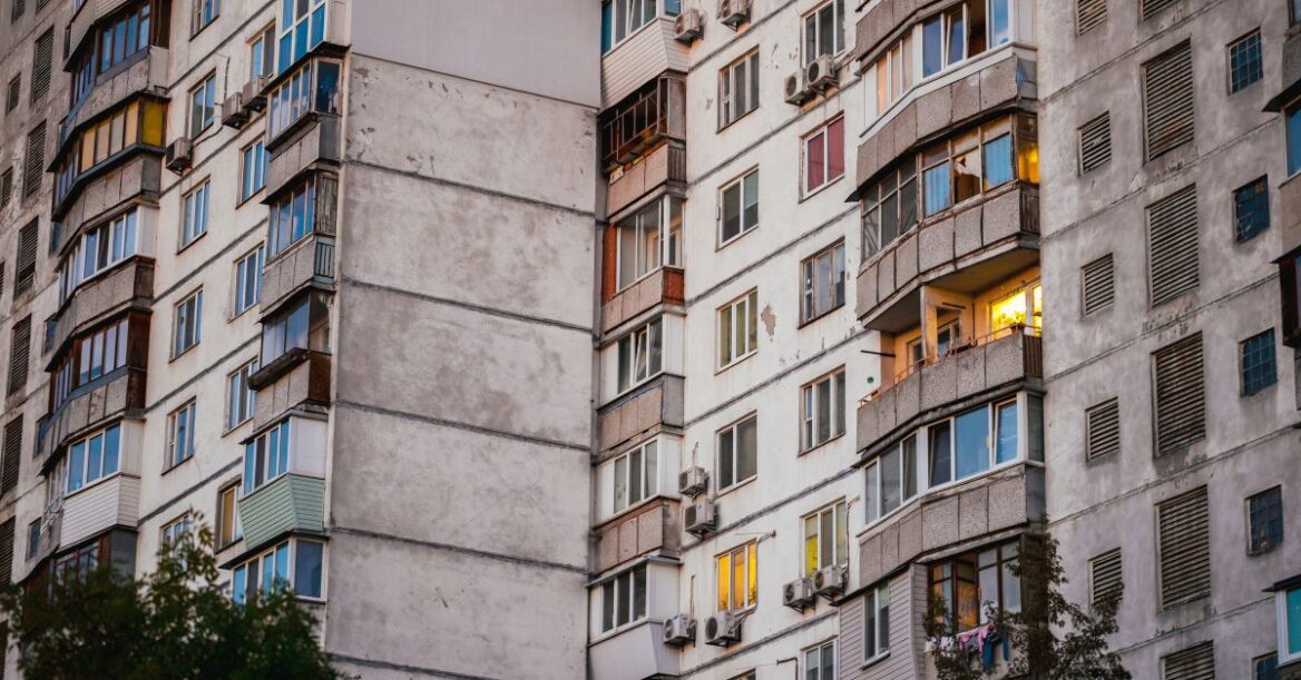 Multi-story concrete apartment building with balconies and windows, some illuminated at dusk, showing signs of age and weathering.