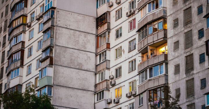 Multi-story concrete apartment building with balconies and windows, some illuminated at dusk, showing signs of age and weathering.