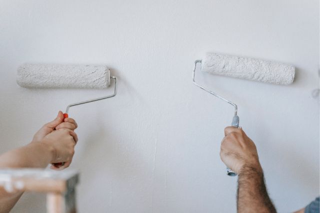 Two hands painting the same wall with roller brushes.
