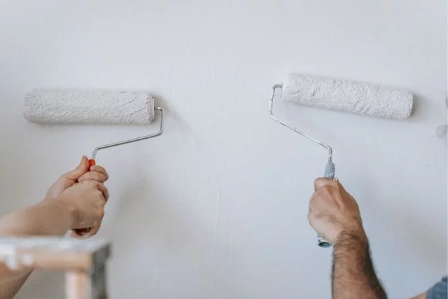 Two hands painting the same wall with roller brushes.