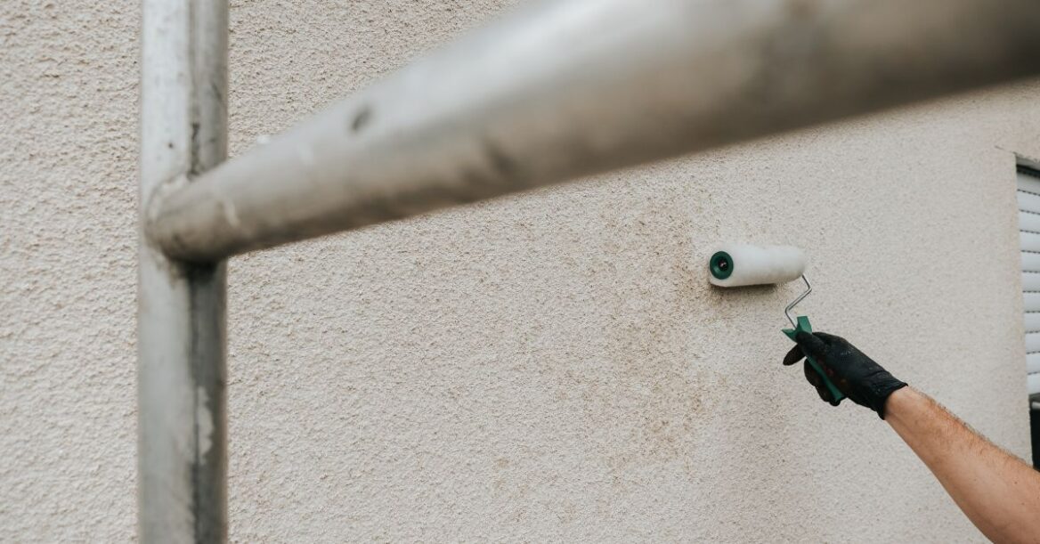 A person wearing a glove uses a paint roller to apply paint to a textured exterior wall, with scaffolding in the foreground.