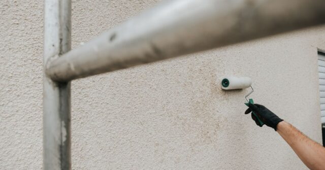 A person wearing a glove uses a paint roller to apply paint to a textured exterior wall, with scaffolding in the foreground.