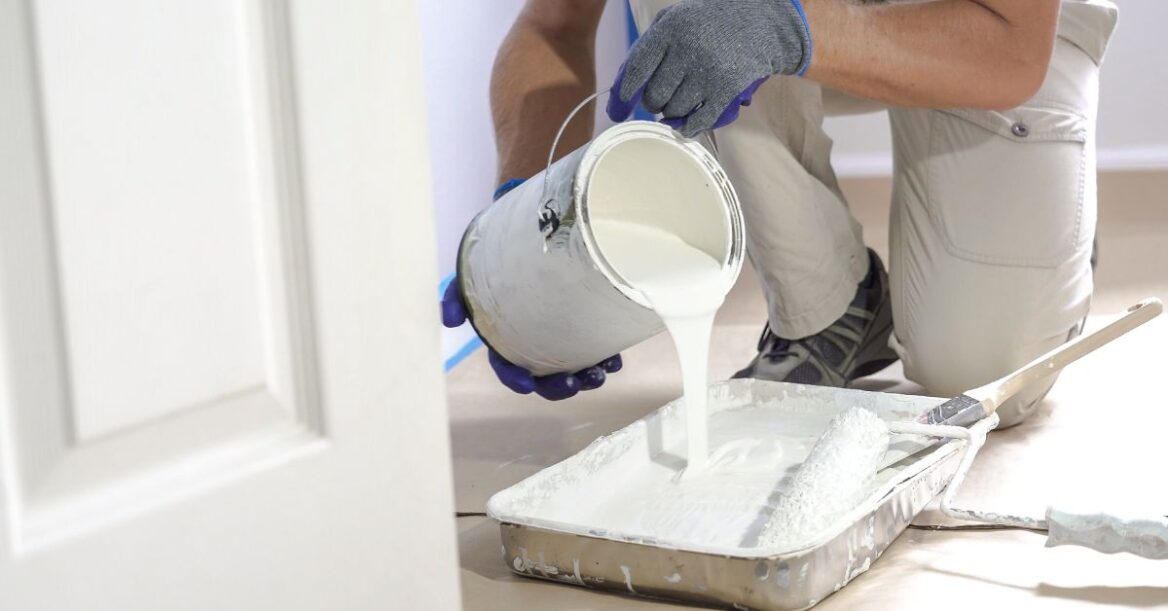 A person wearing gloves and work clothes kneels on the floor while pouring white paint from a bucket into a paint tray, with a roller and brush ready for use.