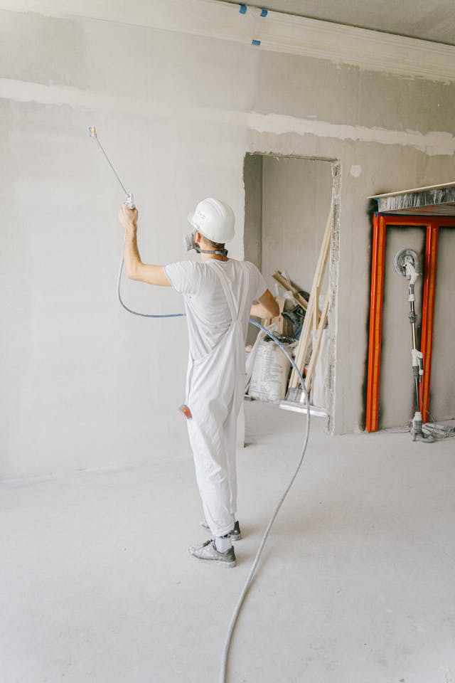 A construction worker in white overalls and a hard hat uses a spray hose to paint an unfinished wall indoors.