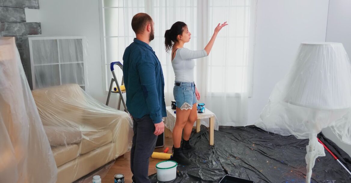 A couple standing in a living room with furniture covered in plastic, discussing wall painting while paint supplies and drop cloths are spread on the floor.