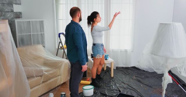 A couple standing in a living room with furniture covered in plastic, discussing wall painting while paint supplies and drop cloths are spread on the floor.