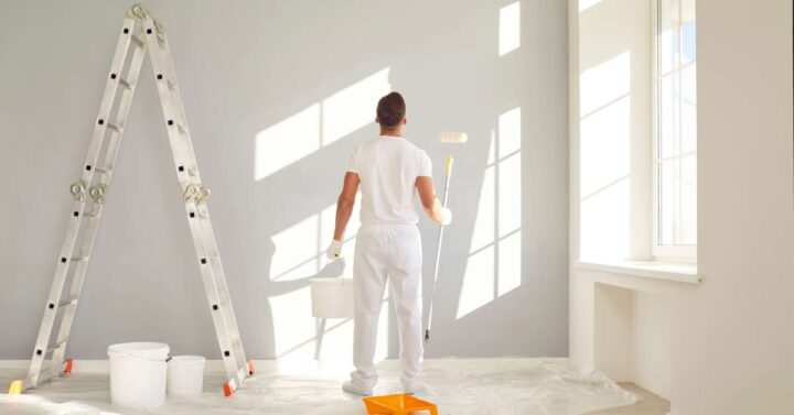 A painter in white overalls uses a roller to paint a light-colored wall in a bright apartment, with a ladder and paint buckets nearby.