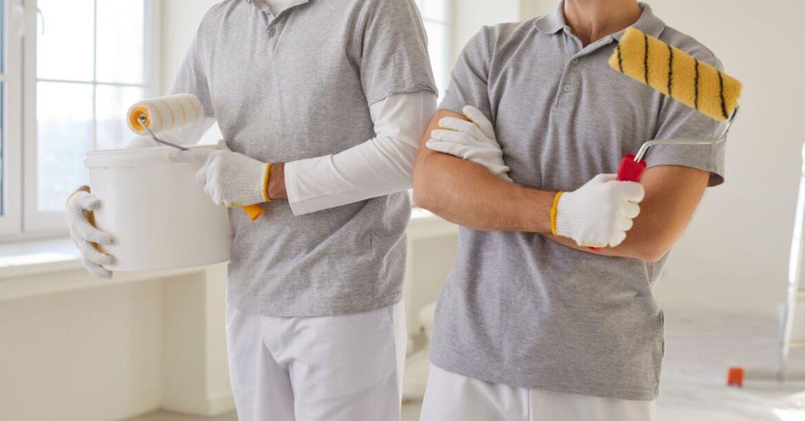 Two professional painters holding paint rollers and a bucket in a bright room ready for interior painting.