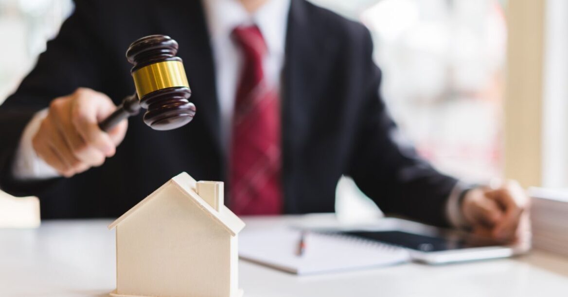 A person in a suit holding a judge’s gavel above a small model house on a desk, symbolizing a property auction or legal decision involving real estate.