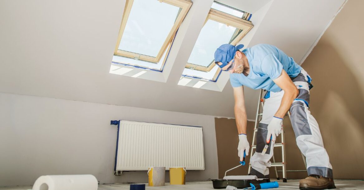 A worker in protective gloves and work clothes uses a paint roller to apply white paint to a wall in a room with skylight windows, with painting supplies scattered on the floor nearby.