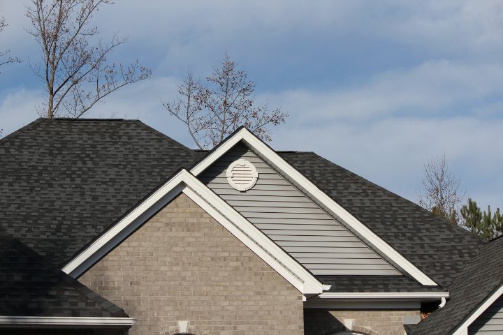 Close-up view of a residential house with asphalt shingle roofing and gable vent, featuring gray vinyl siding and brick exterior under a clear blue sky.