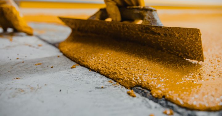 Close-up of a gloved hand using a metal trowel to spread epoxy coating across a concrete floor during industrial flooring installation.