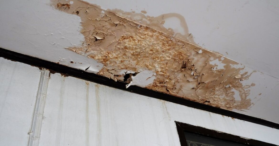 Close-up of a ceiling with severe water damage, showing peeling paint, cracks, and brown stains above an interior wall and window frame