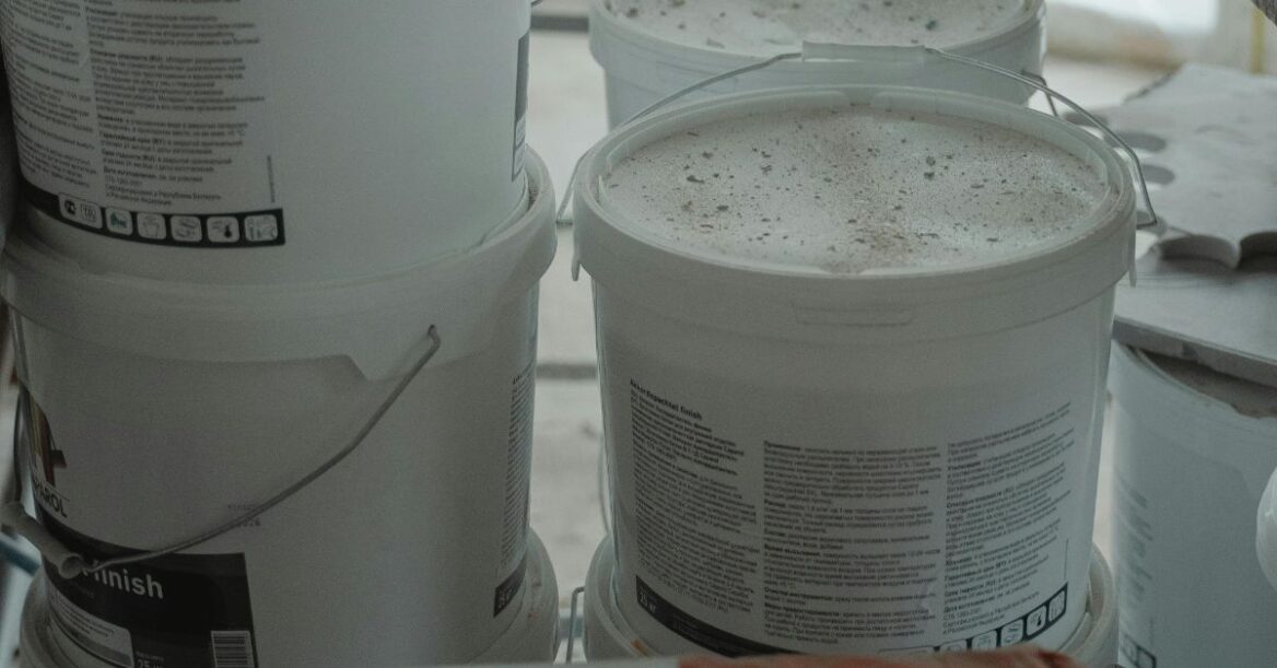 Close-up of several white plastic buckets with printed labels, covered in dust and stacked in a workshop or construction area.