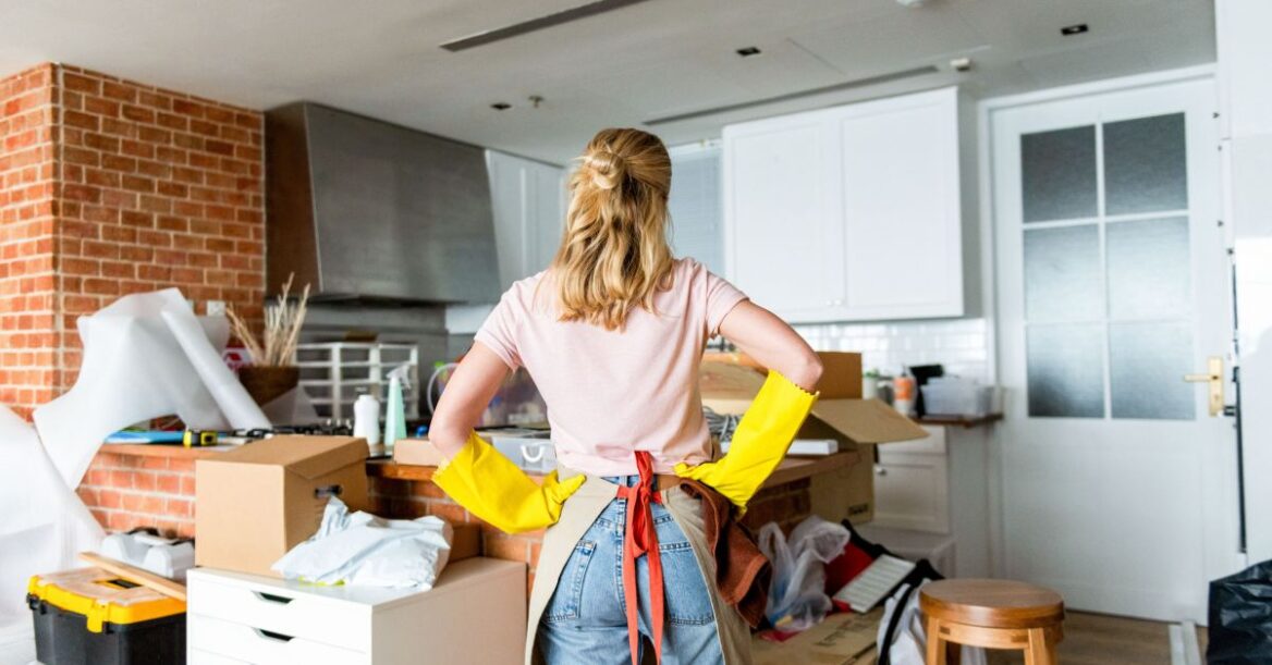 Woman wearing cleaning gloves stands in a cluttered room surrounded by boxes and supplies.