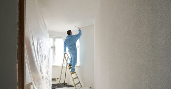 Person in protective clothing standing on a ladder and painting a white interior wall in a room covered with plastic sheeting during renovation.