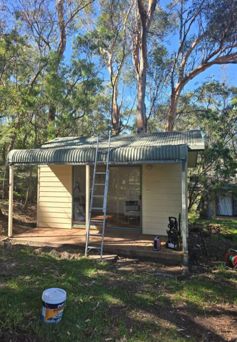 A small beige-colored cabin with a weathered corrugated roof and sliding glass doors before repainting and restoration.