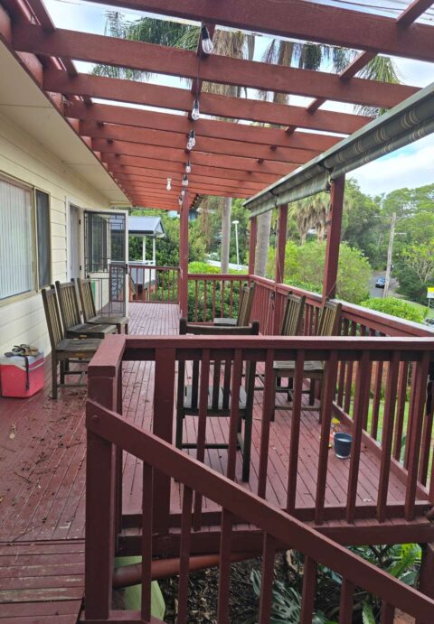 An older red-stained wooden deck with chairs and a pergola, showing weathered timber before repainting.