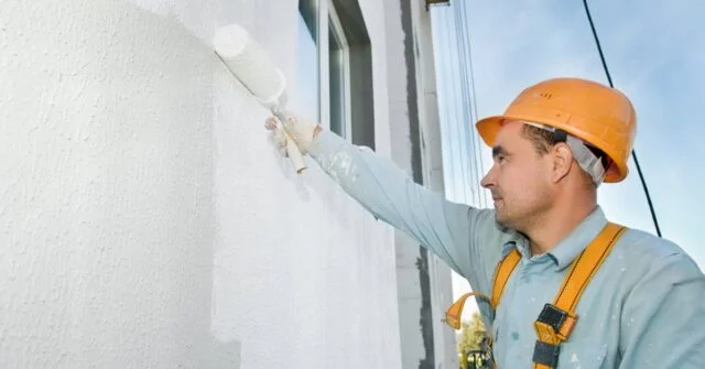 A professional painting contractor in a hard hat painting an exterior wall of a house.