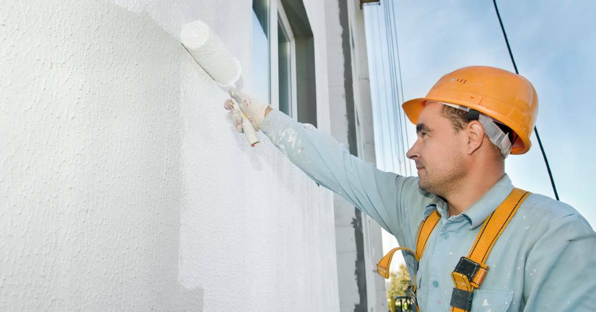 A professional painting contractor in a hard hat painting an exterior wall of a house.