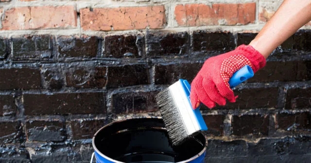 Person wearing a red glove and holding a blue paintbrush is painting black primer on a brick wall.