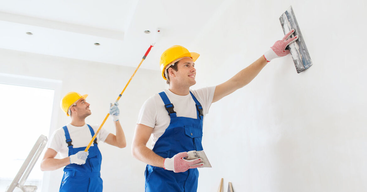 Two professional painters in blue overalls with yellow hard hats, painting a room white.