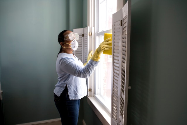 Lady wearing a mask, goggles and yellow gloves is cleaning a window with a sponge and cleaning solution.