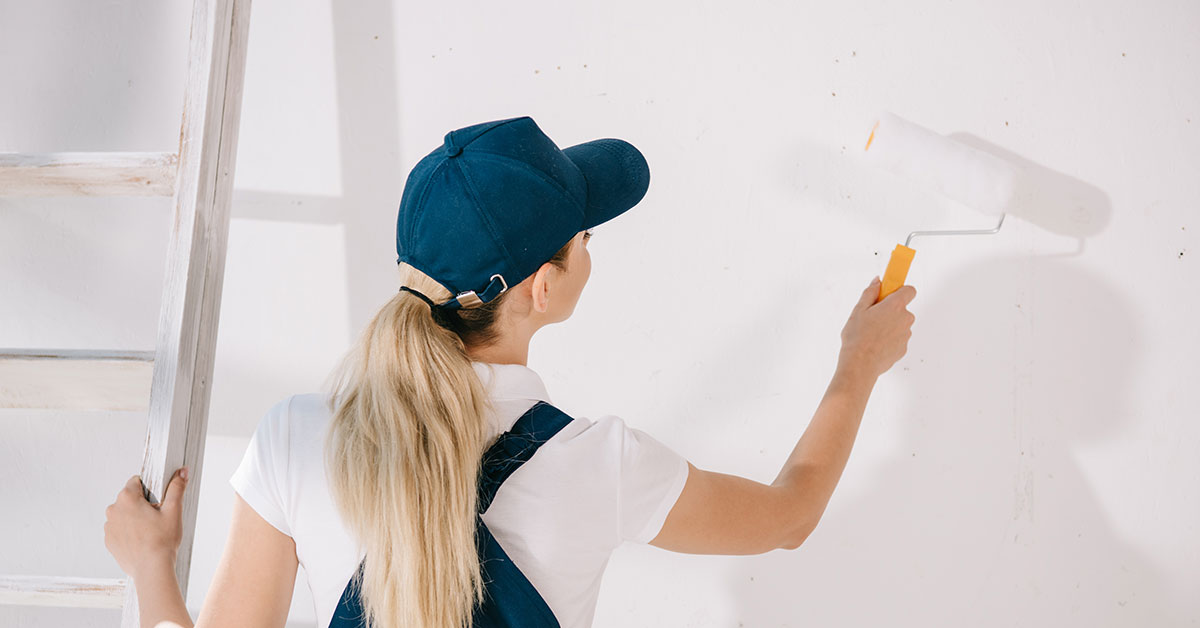Women holding a ladder in one hand and painting a wall with a roller in the other.